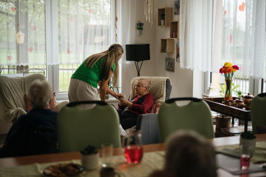 Cuidadora conversando com uma senhora sentada numa poltrona, numa sala de convivência