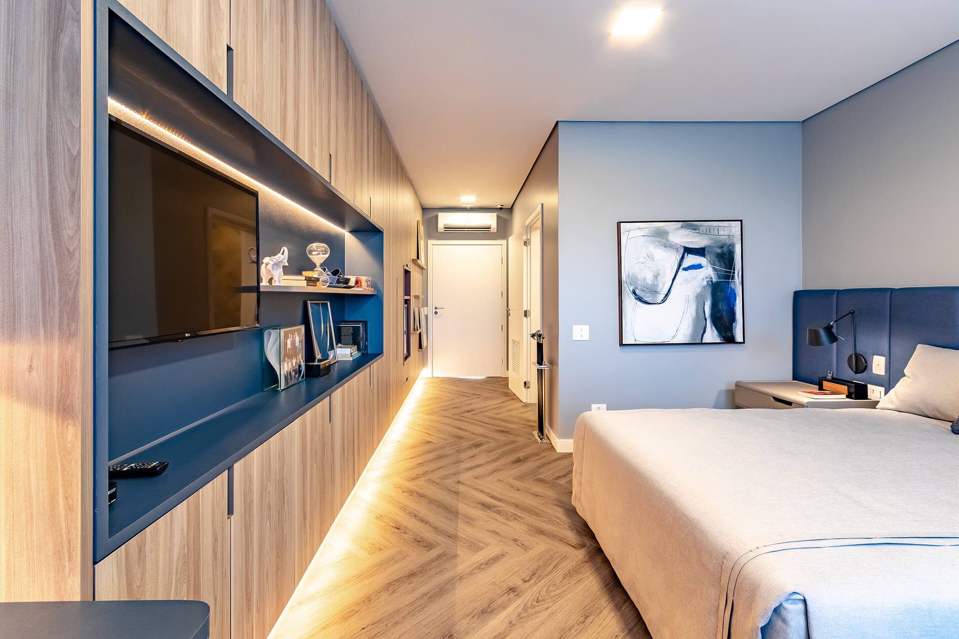 Suíte com cama de casal, painel de madeira com TV e piso de madeira em espinha de peixe